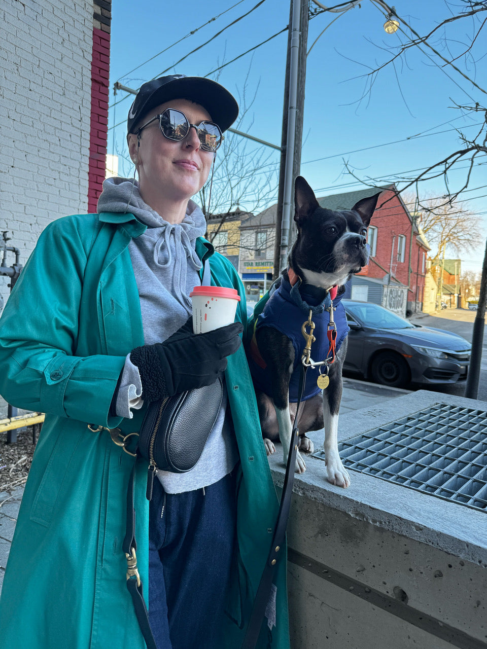Caucasian female wearing sunglasses and holding a coffee looks upwards and away from the camera. To her right is a Boston terrier perched up on a cement block in a public space. 