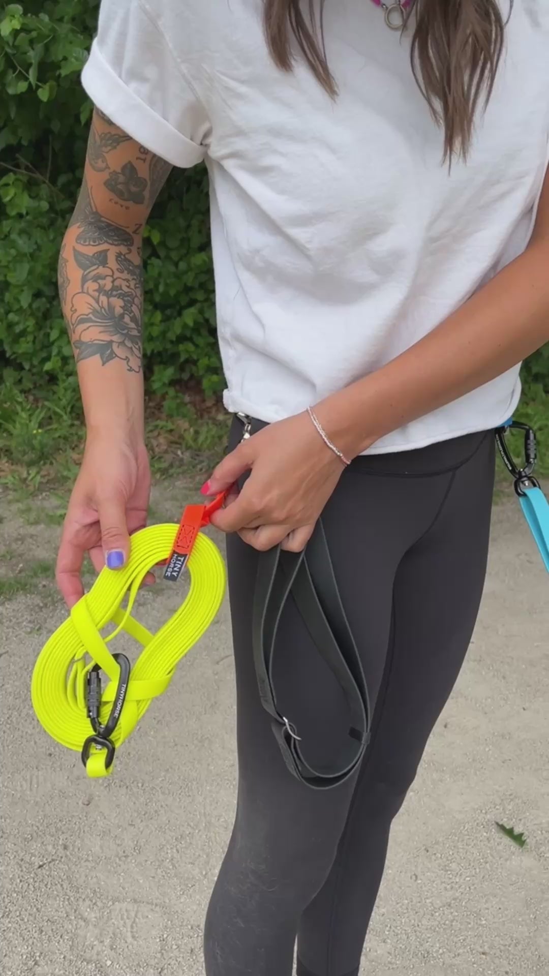 Female, Caucasian dog walker demonstrates how the Keeper can be attached and detached from the Leader walking belt. The Keeper is looped through a trainer long line. The scene takes place in a park during the summer time.